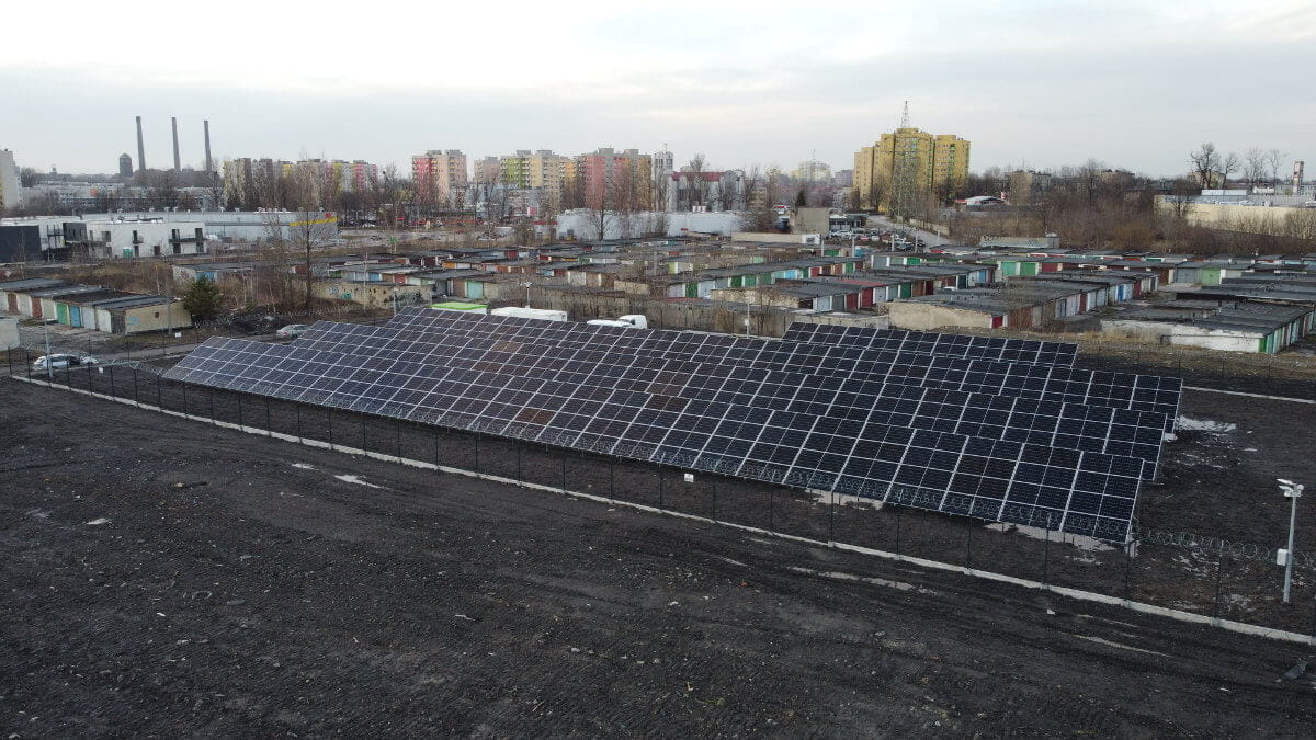 Gruntowa farma fotowoltaiczna mikrosieci, widziana od strony południowej. W tle widoczne zabudowania miejskie (bloki, garaże).