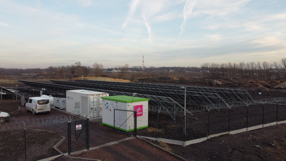 Ogrodzony teren mikrosieci, a na nim: pomieszczenie techniczne, magazyn energii, agregat gazowy wraz ze zbiornikiem paliwa. Obok nich stoi samochód dostawczy. Za nimi gruntowa farma fotowoltaiczna, widziana od strony płn.-zach. (od tyłu). 