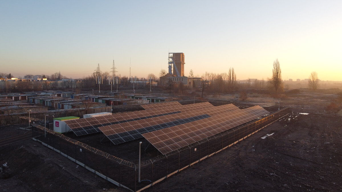 Obiekty mikrosieci widziane o wschodzie słońca: gruntowa farma fotowoltaiczna, za nią pomieszczenie techniczne i magazyn energii, w tle mikroturbiny wiatrowe, szyb d. kopalni Szombierki i zabudowania miejskie.