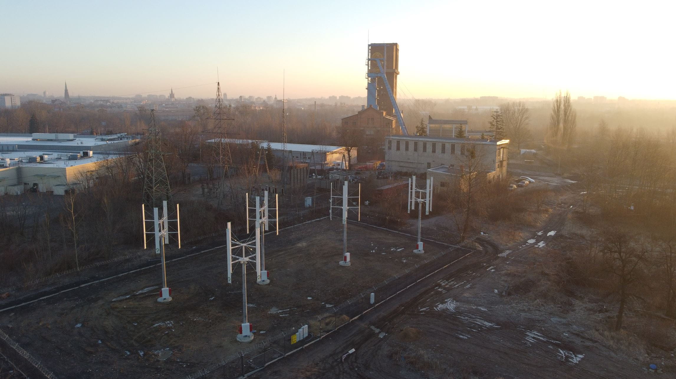 Widok z lotu drona o wschodzie słońca: mikroturbiny wiatrowe na ogrodzonym gruncie, w tle szyb d. kopalni Szombierki, zabudowania przemysłowe i panorama Bytomia. 