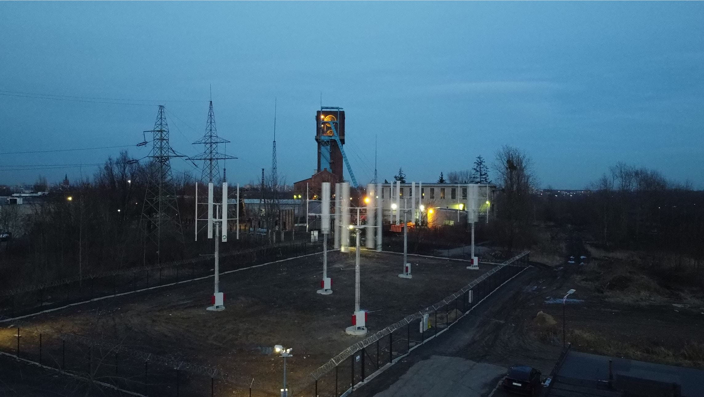 Mikroturbiny wiatrowe (rozmyte z powodu ruchu wirników) widziane w nocy; wokół gruntu ogrodzenie, w tle szyb d. kopalni Szombierki, oświetlone zabudowania przemysłowe i słupy energetyczne.