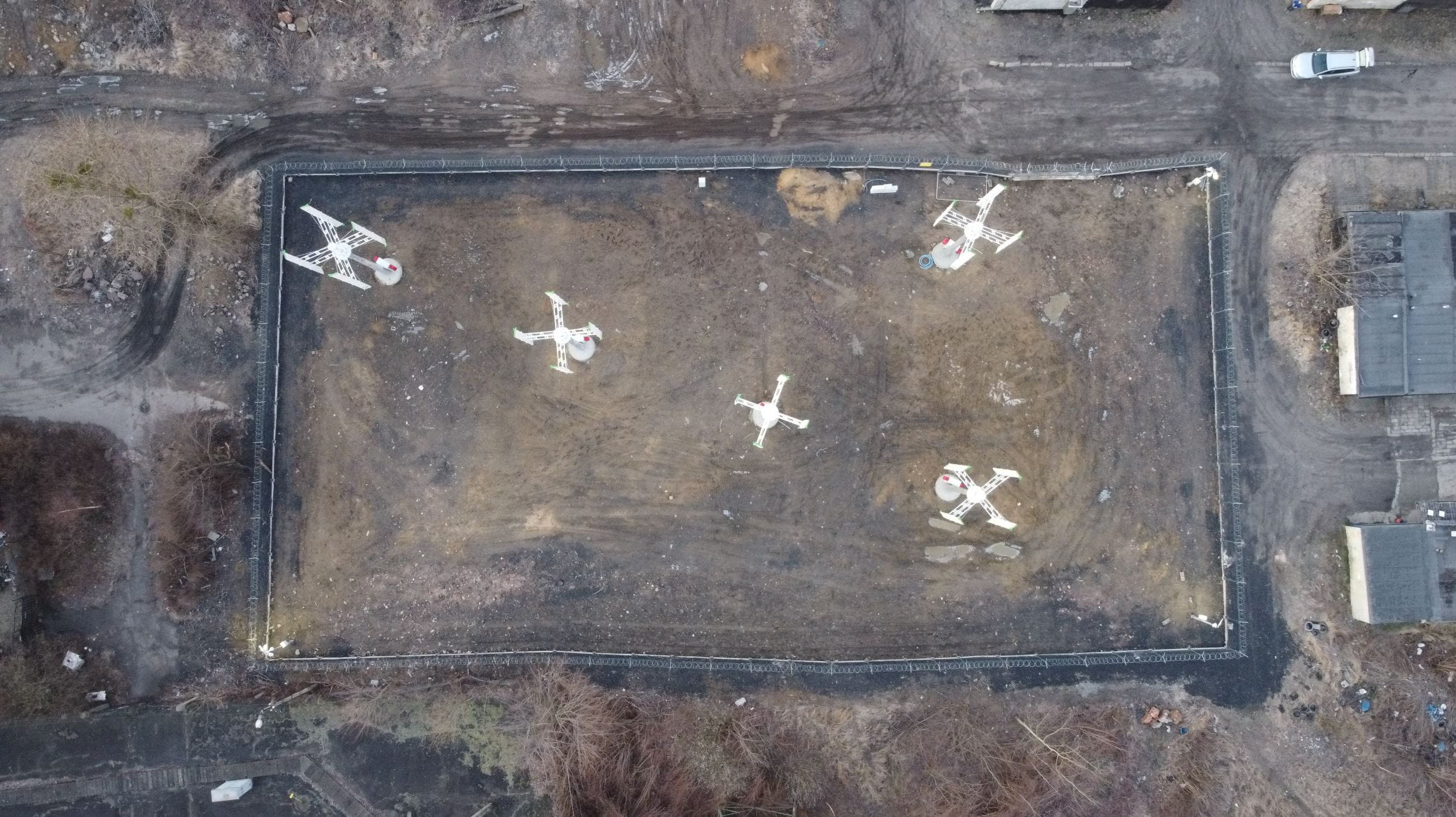 Widok z góry na obracające się mikroturbiny wiatrowe o pionowej osi obrotu, ogrodzone i posadowione na zrekultywowanym ternie poprzemysłowym.