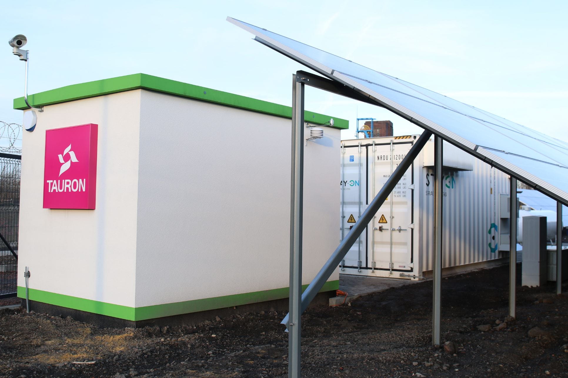 Biały, murowany budynek z zielonym cokołem i dachem oraz dużym logotypem TAURON (w kolorze magenta); obok fragment gruntowej farmy fotowoltaicznej i kontener magazynu energii.