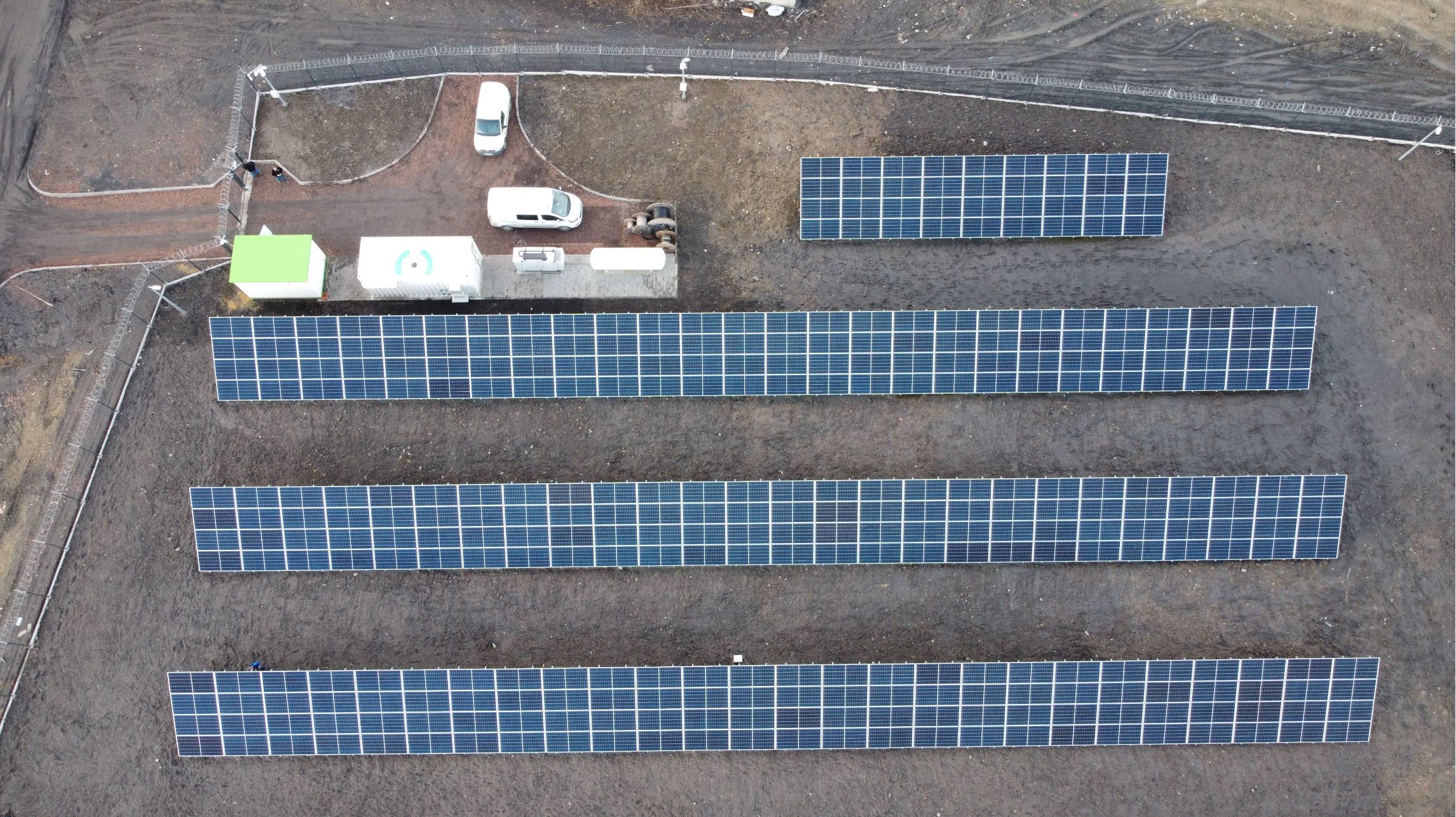 Widok z góry na gruntową farmę fotowoltaiczną, pomieszczenie techniczne, kontener magazynu energii, agregat gazowy ze zbiornikiem paliwa; teren wokół ogrodzony i posadowiony na gruncie poprzemysłowym 