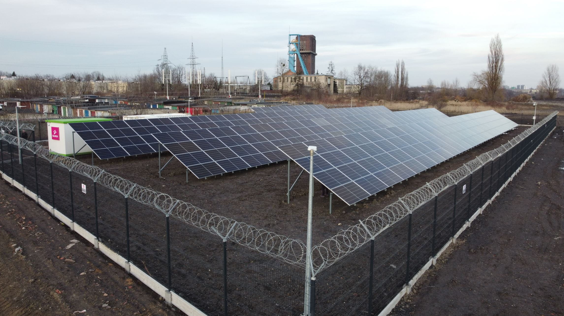Widok z narożnika działki mikrosieci: gruntowa farma fotowoltaiczna, za nią kontener techniczny i magazynu energii, wokół obiektów ogrodzenie i kamery przemysłowe; w tle mikroturbiny wiatrowe, szyb kopalniany i zabudowania przemysłowe.