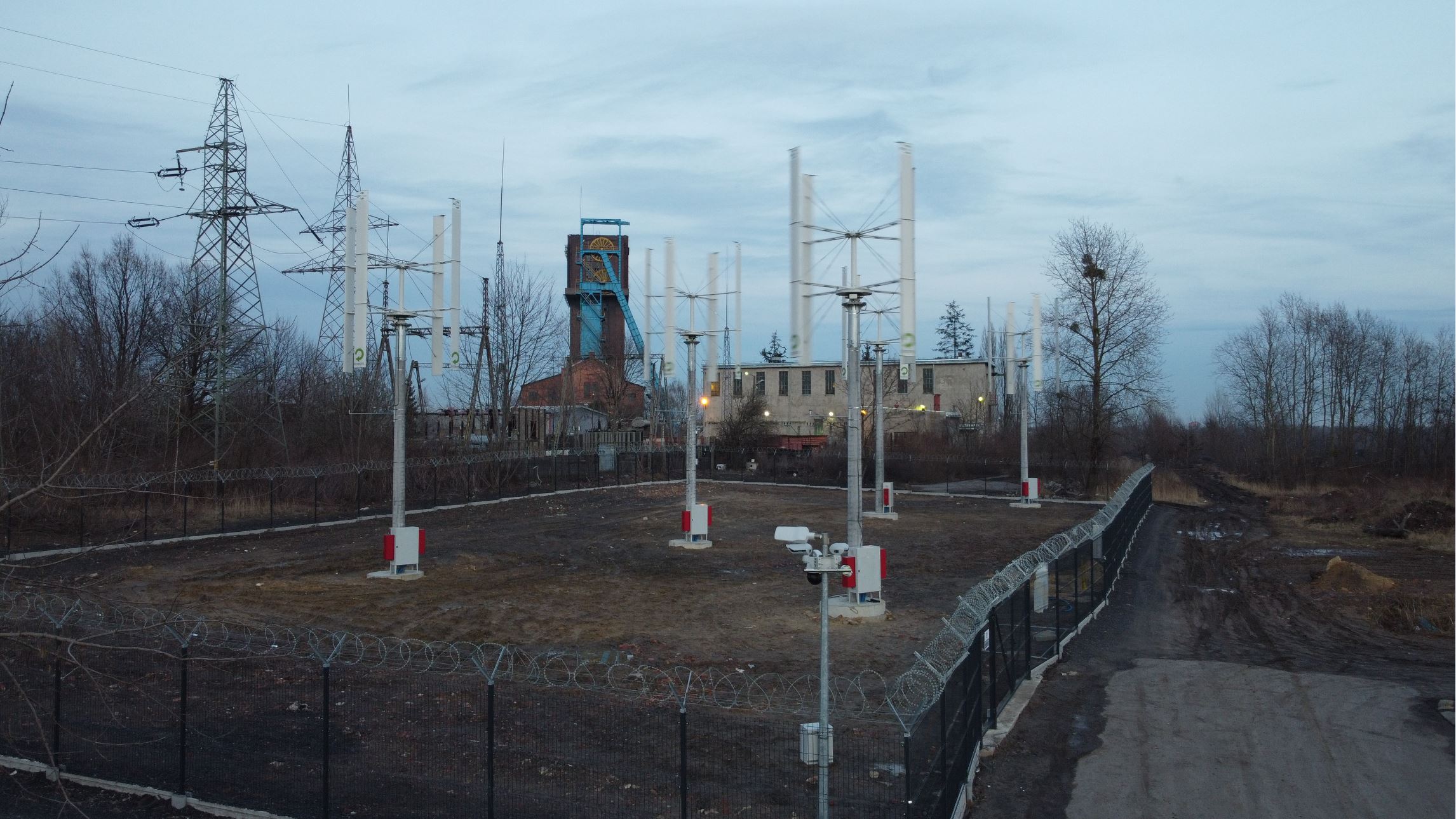 Pięć mikroturbin wiatrowych o pionowej osi obrotu, wokół przemysłowe ogrodzenie i monitoring; w tle szyb kopalni, słupy energetyczne i zabudowania przemysłowe.